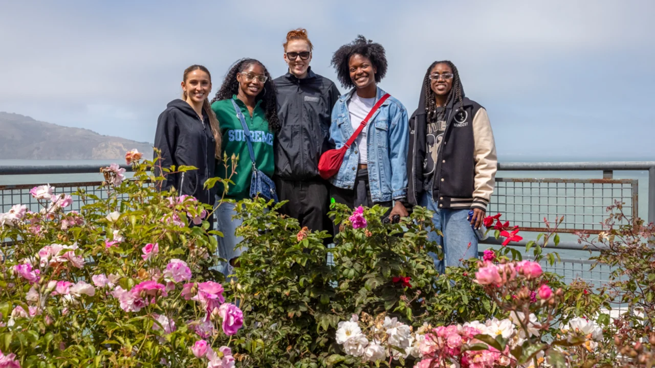 Five individuals from the Golden State Valkyries team pose together behind a vibrant bed of pink and white roses, with a scenic coastal backdrop featuring water and distant hills under a partly cloudy sky.