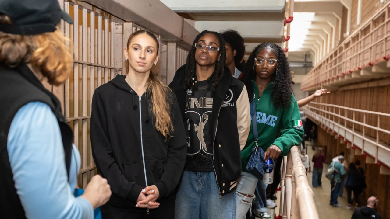 Three individuals stand in a long corridor lined with barred cells, observing their surroundings as a parks conservancy Alcatraz tour guide gestures nearby.