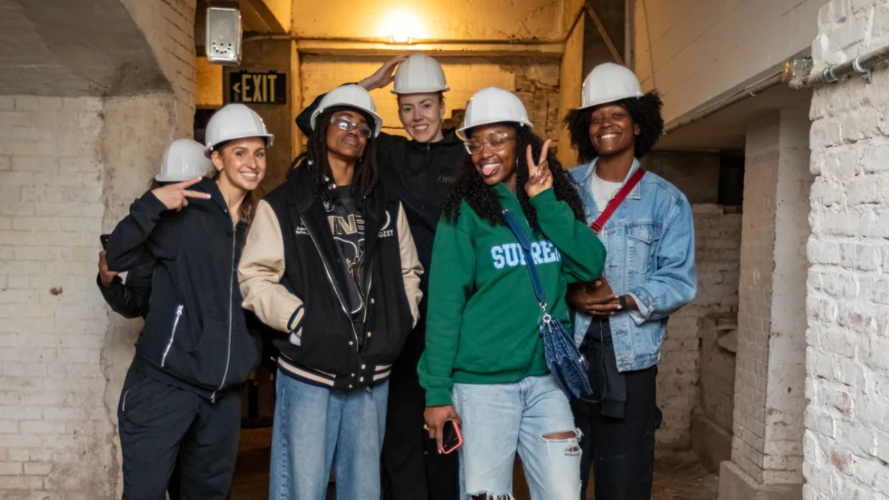 A group of five individuals wearing hard hats pose cheerfully in a narrow, whitewashed brick corridor with an illuminated exit sign.