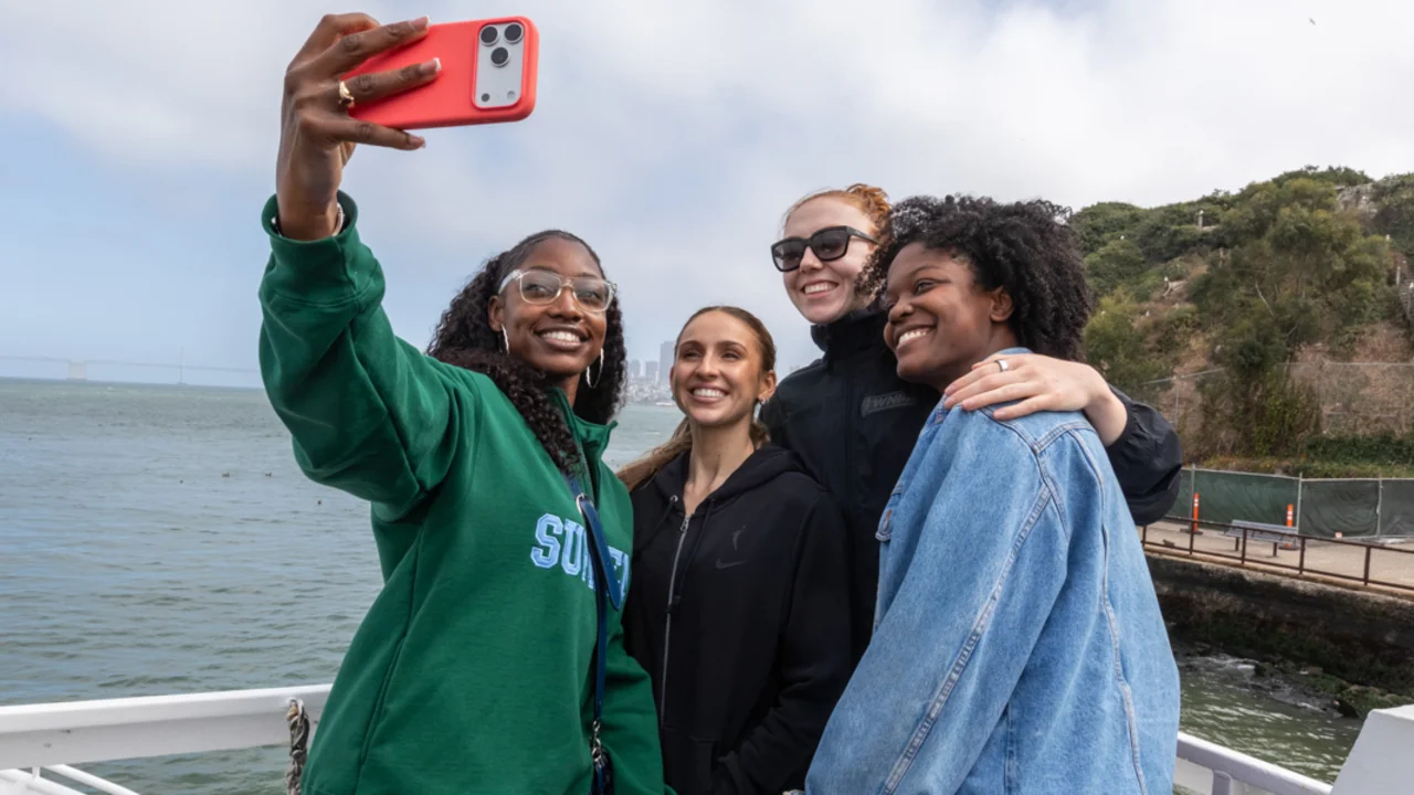 Four individuals take a selfie together on a boat, smiling with a waterfront and distant city skyline in the background.