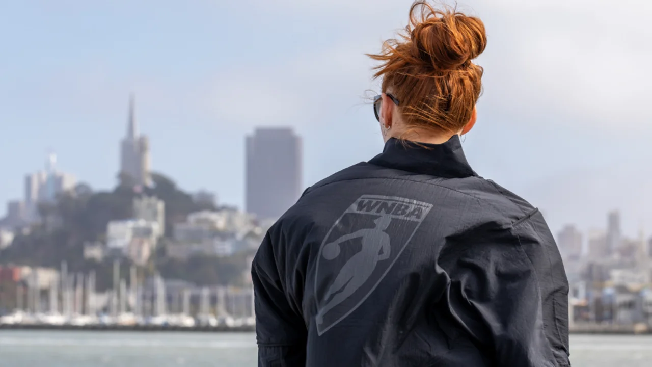 A person with red hair in a bun, wearing a dark jacket with a WNBA logo, stands by the water facing a city skyline.