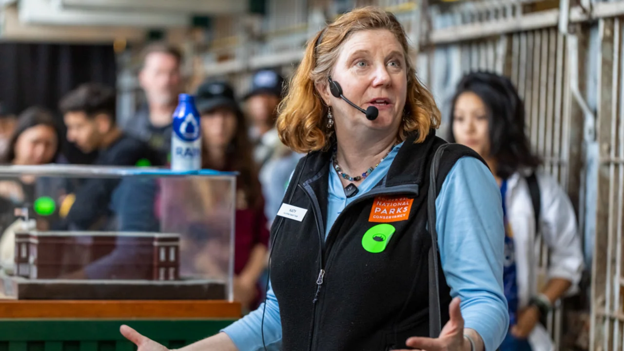 A guide wearing a headset and a Golden Gate National Parks Conservancy vest gestures while speaking to a group during a tour.