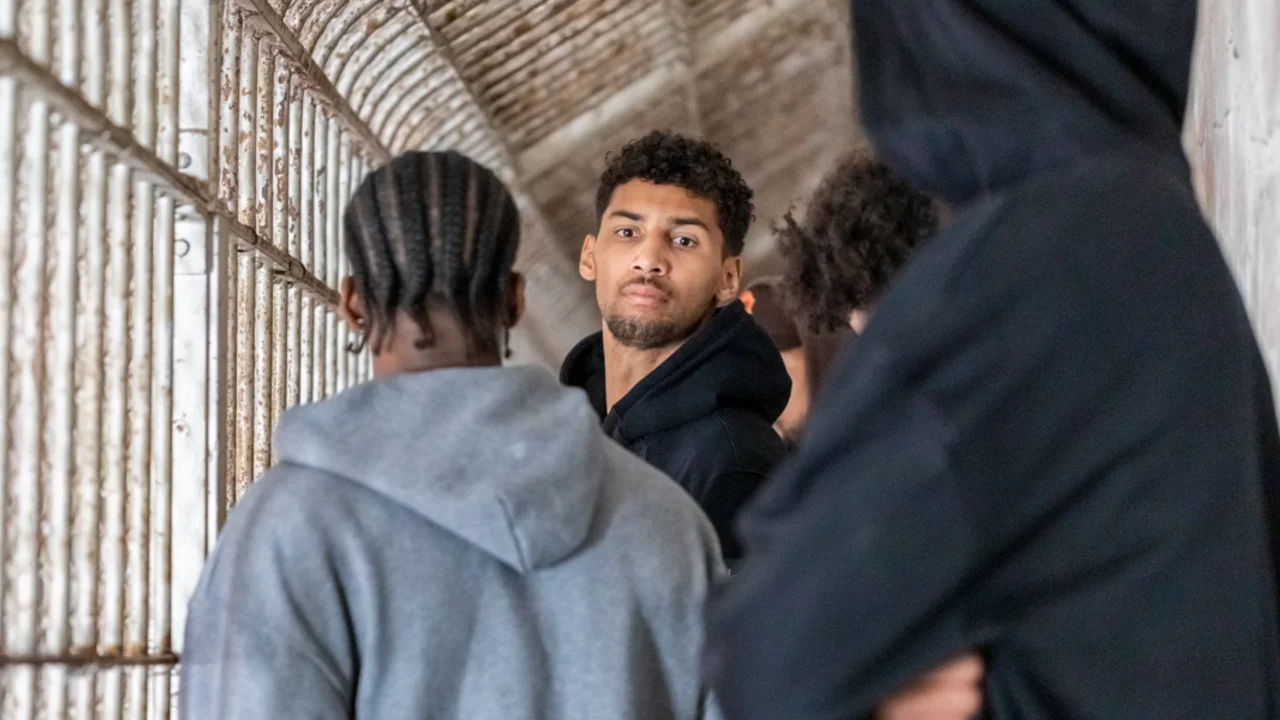 A group of players from the Golden State Warriors stand in a narrow, barred corridor on Alcatraz Island, with one person looking directly at the camera.