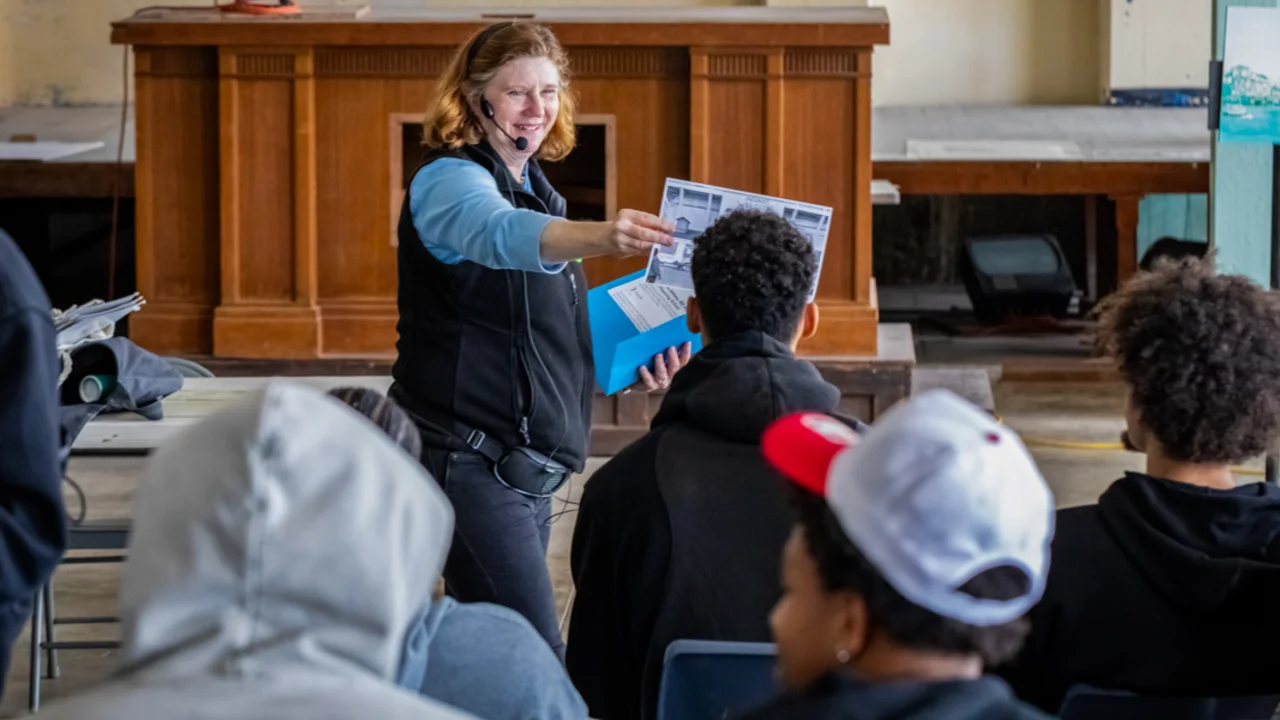 A Alcatraz Custom Tour guide stands before a group from the Golden State Warriors, holding up a printed document while speaking into a headset microphone in a room with wooden furnishings.