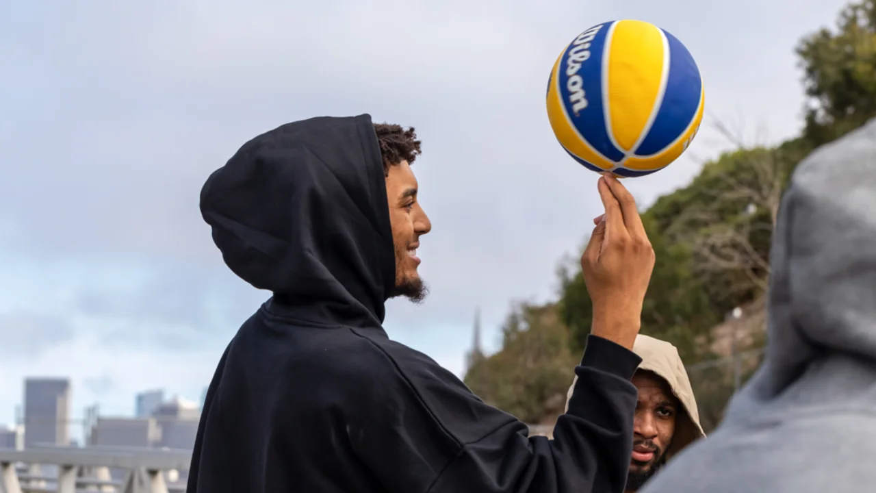 A player from the Golden State Warriors in a black hoodie balances a yellow and blue volleyball on their finger while smiling, with others nearby and a San Francisco city skyline in the background.