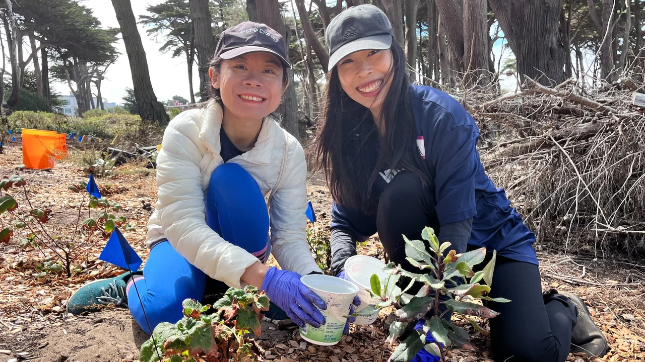 Two volunteers kneel and smile for the camera while collecting weeds.
