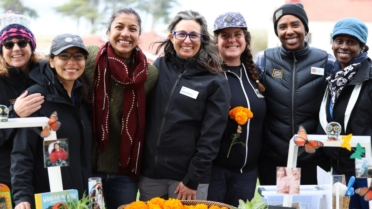 A group of people smiling, bright orange marigolds, photos, butterfly art, and other effects are present at the table before them in celebration of Diwali and Dia de los Muertos.