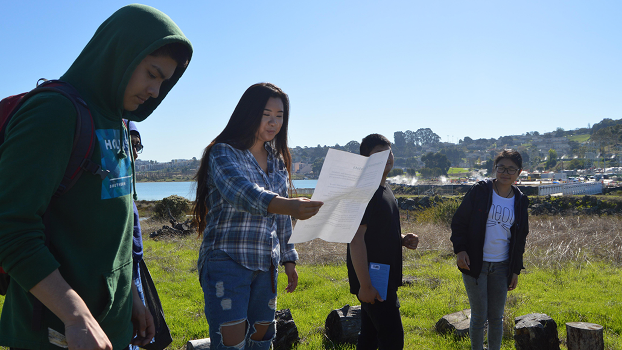 A group of four high school students stand in a line outdoors while leading a presentation about the natural environment 