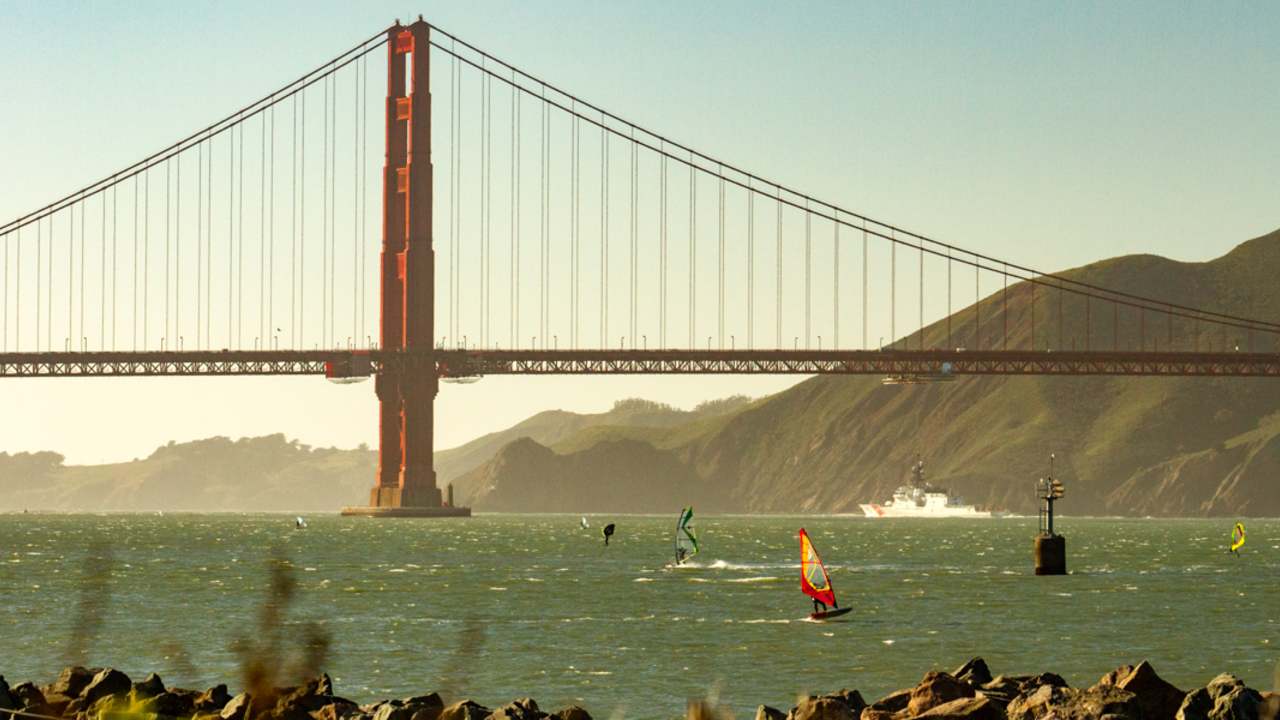 Windsurfers cruise across bay waters with the Golden Gate Bridge spanned across the background.