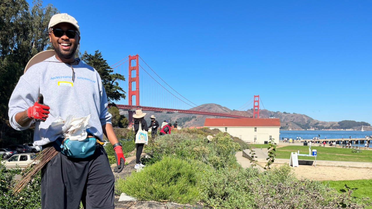 A volunteer gives a thumbs up at Crissy Field in San Francisco with a backdrop of the Golden Gate Bridge and the Warming Hut.