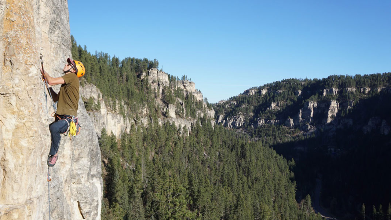 Daniel-Joannes_2x1.jpg A person scales a rock wall outdoors overlooking a forest.