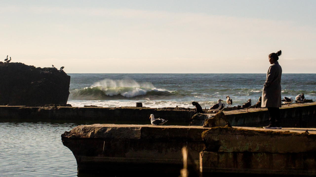 A park visitor looks out across the Sutro Bath ruins at Lands End. A wave breaks alongside the ocean rocks and birds in the background.