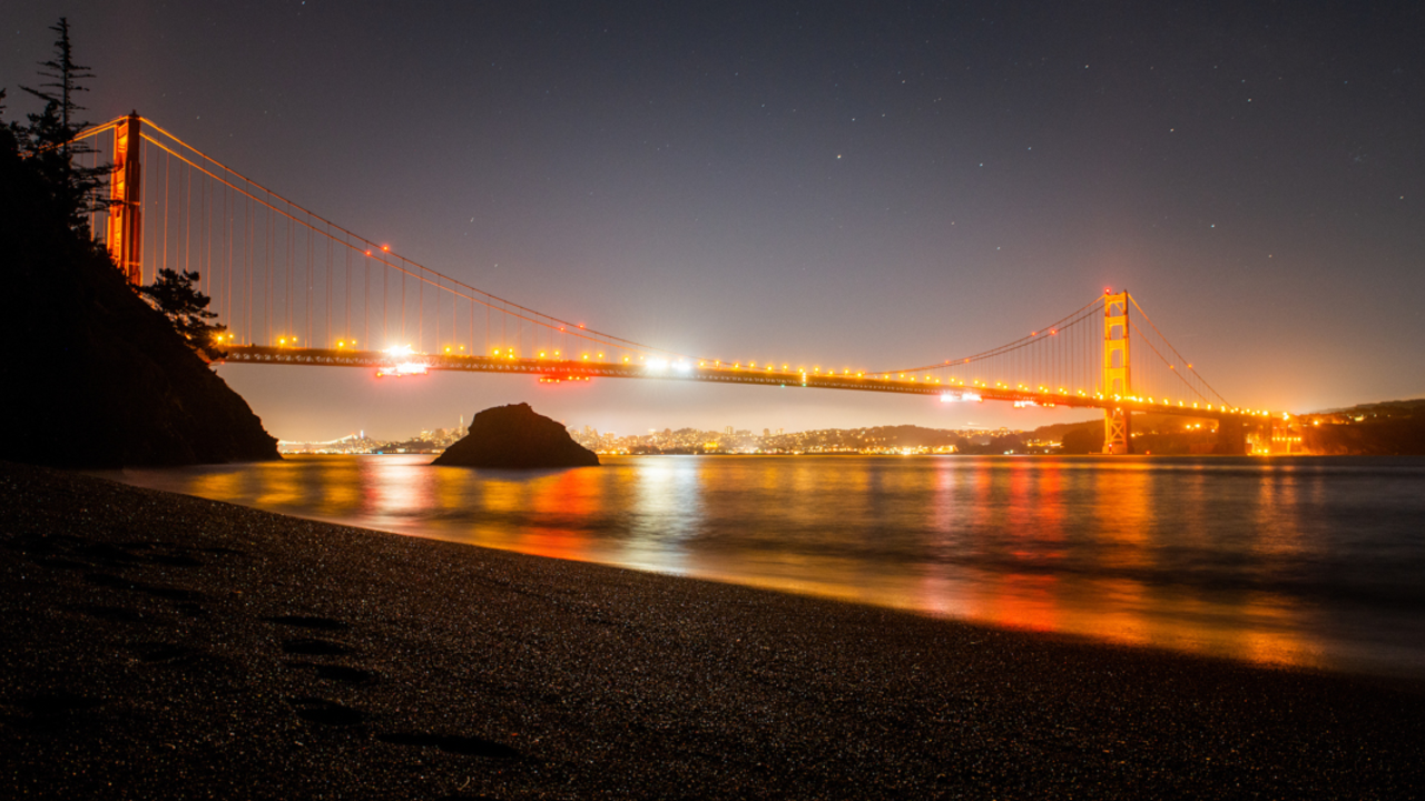 A night view of the Golden Gate Bridge from Kirby Cove. The lights from the bridge glow on the water and there are stars twinkling overhead.