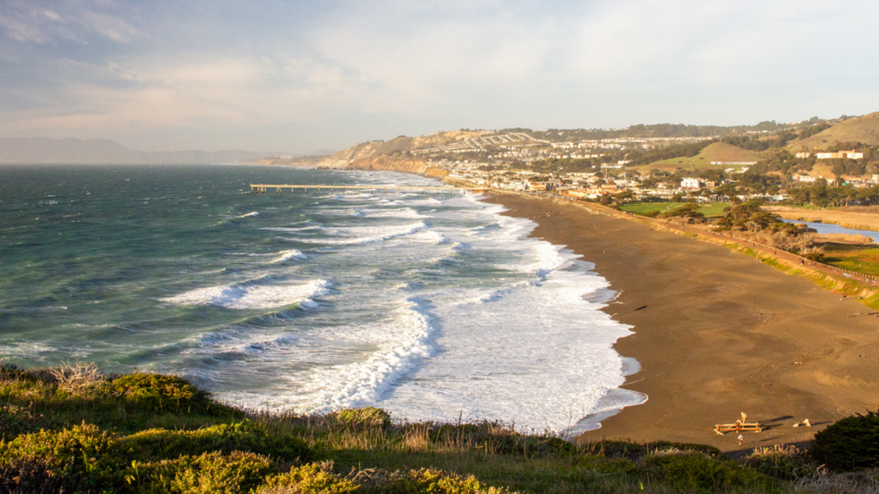 An expansive view of the coastline in Pacifica, CA from Mori Point. A long fishing pier stands out over the beach.