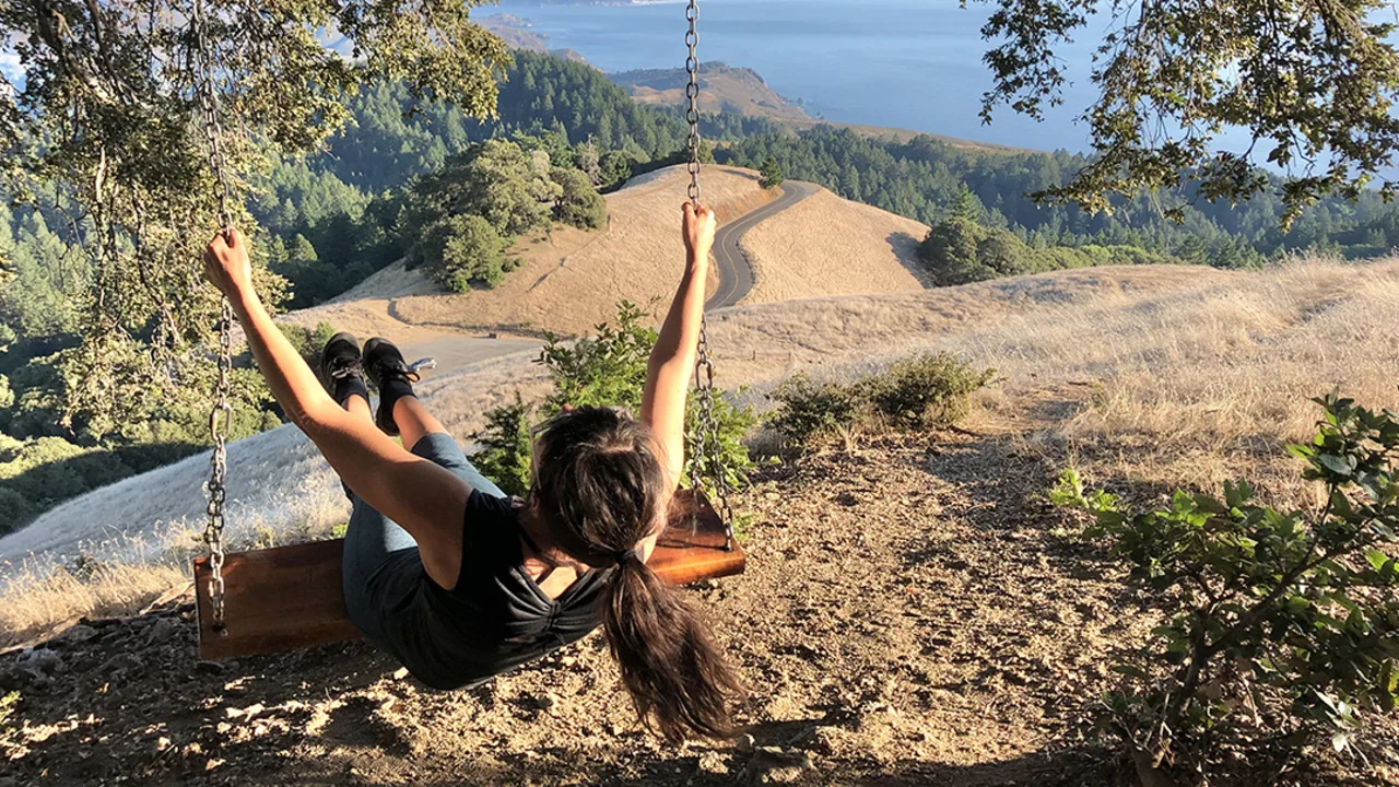 A person swings from a tree with a view of Mt. Tam and the Pacific Ocean.