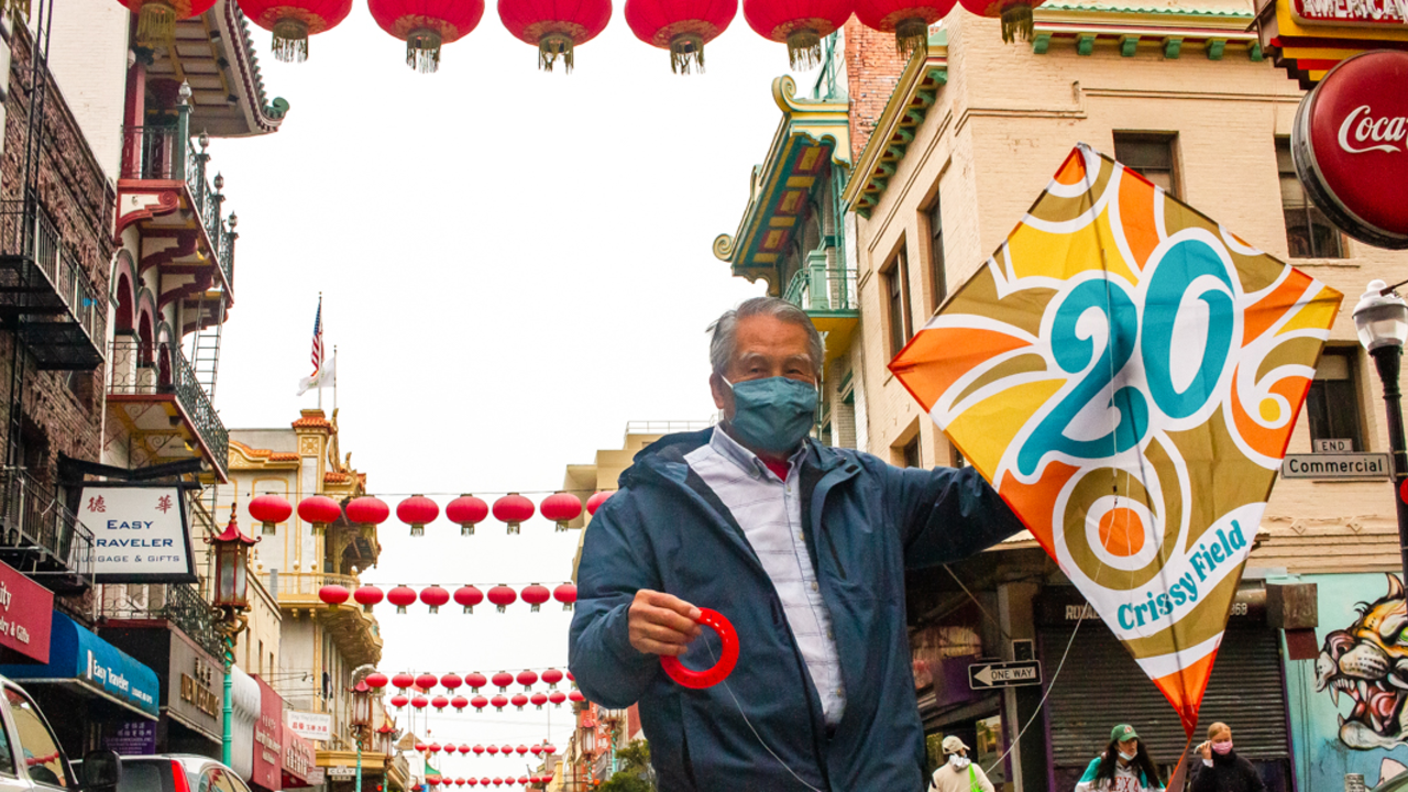 An elderly man holds up a commemorative kite that reads "20 Crissy Field" in San Francisco's famous Chinatown neighborhood.