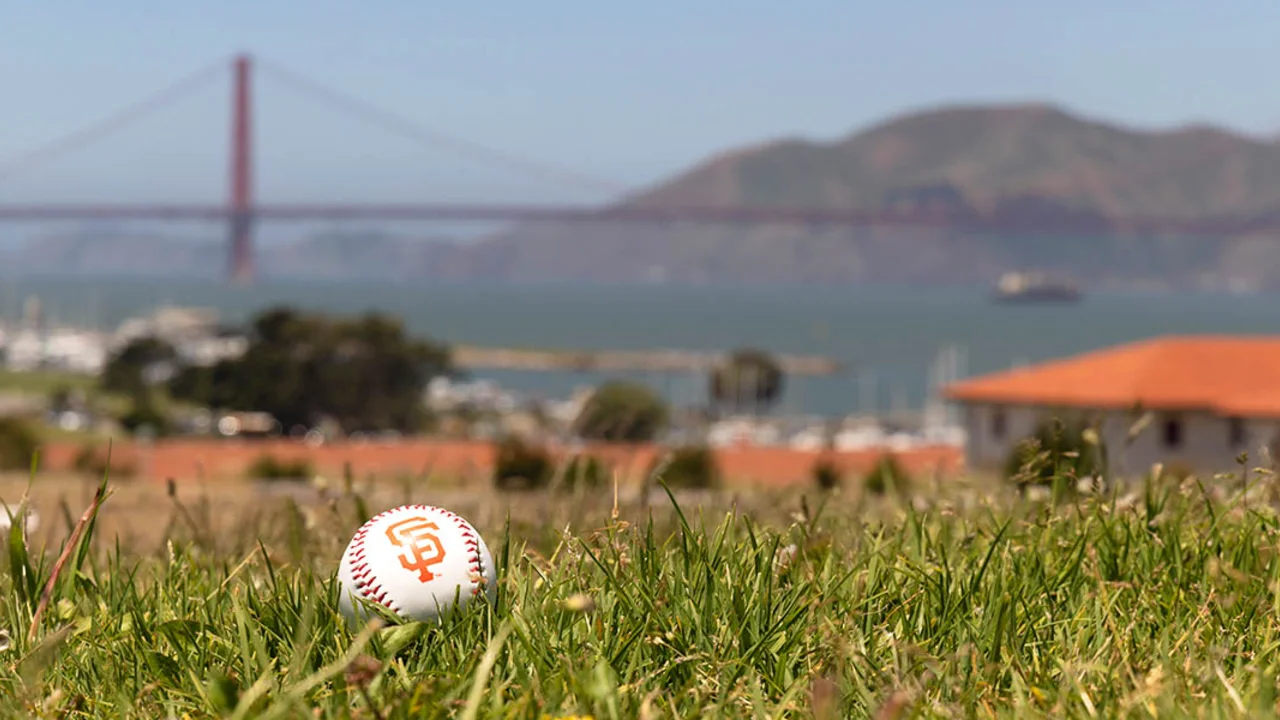 A baseball with the SF Giants logo lays on the green lawn at Fort Mason with a view of the Golden Gate Bridge in the background.
