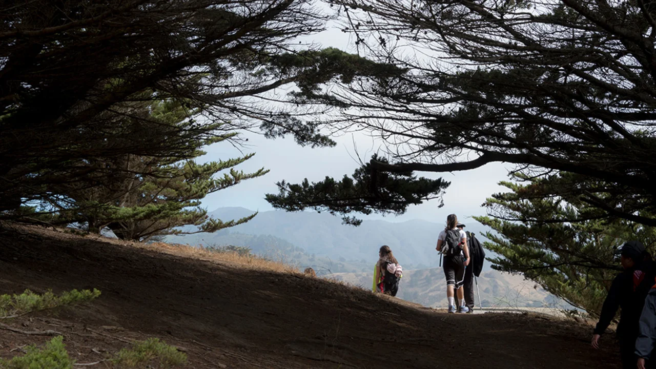 People hiking down a winding path through trees at Rancho Corral de Tierra, sloping hillsides are visible ahead and in the background.