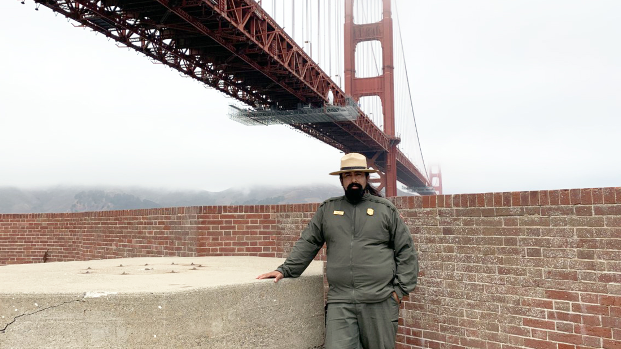 NPS Ranger Erick Cortes stands in front of the Golden Gate Bridge at Fort Point National Historic Site.