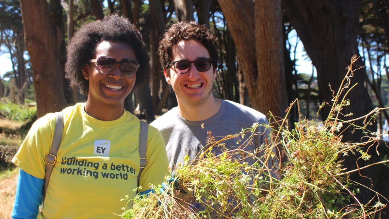 two people smile for the camera while holding plants they removed during a volunteer event