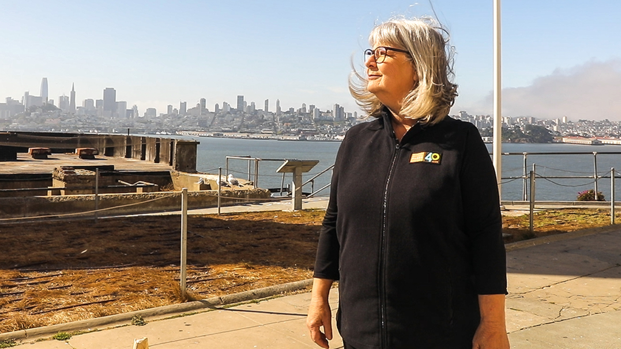 Nicki Phelps stands on Alcatraz Island with a view of San Francisco behind her.