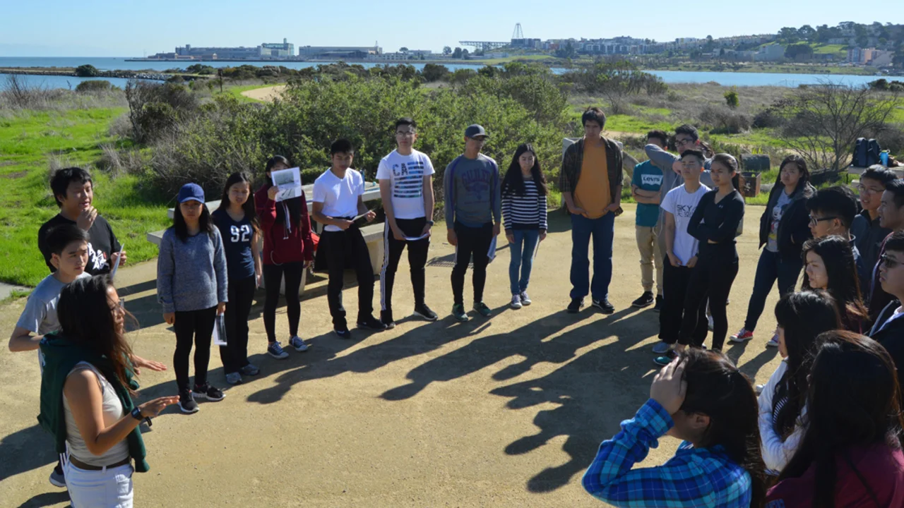 High school students stand in a circle outdoors while listening to an educator describe the natural environment surrounding them