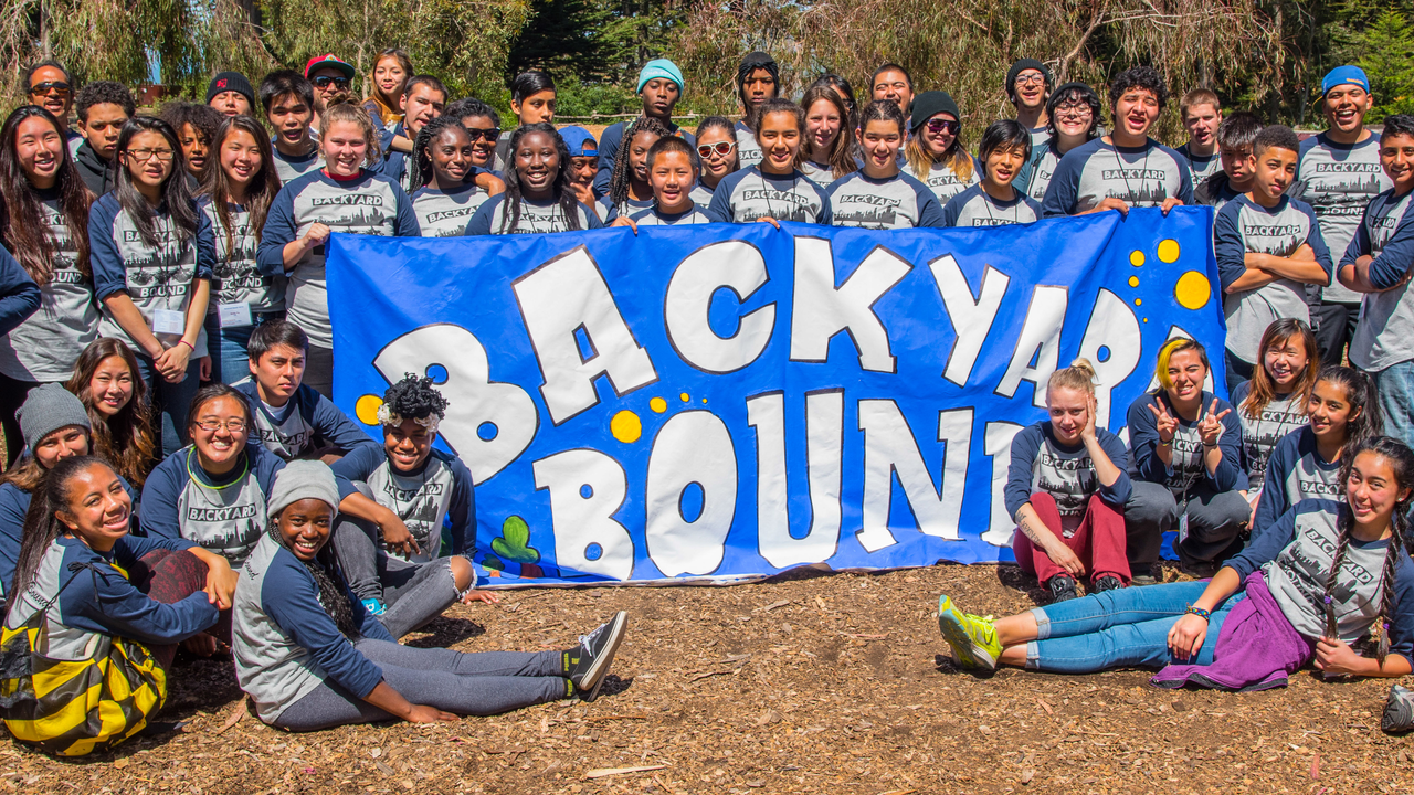 A large group of youth and youth workers gather around a banner reading Backyard Bound