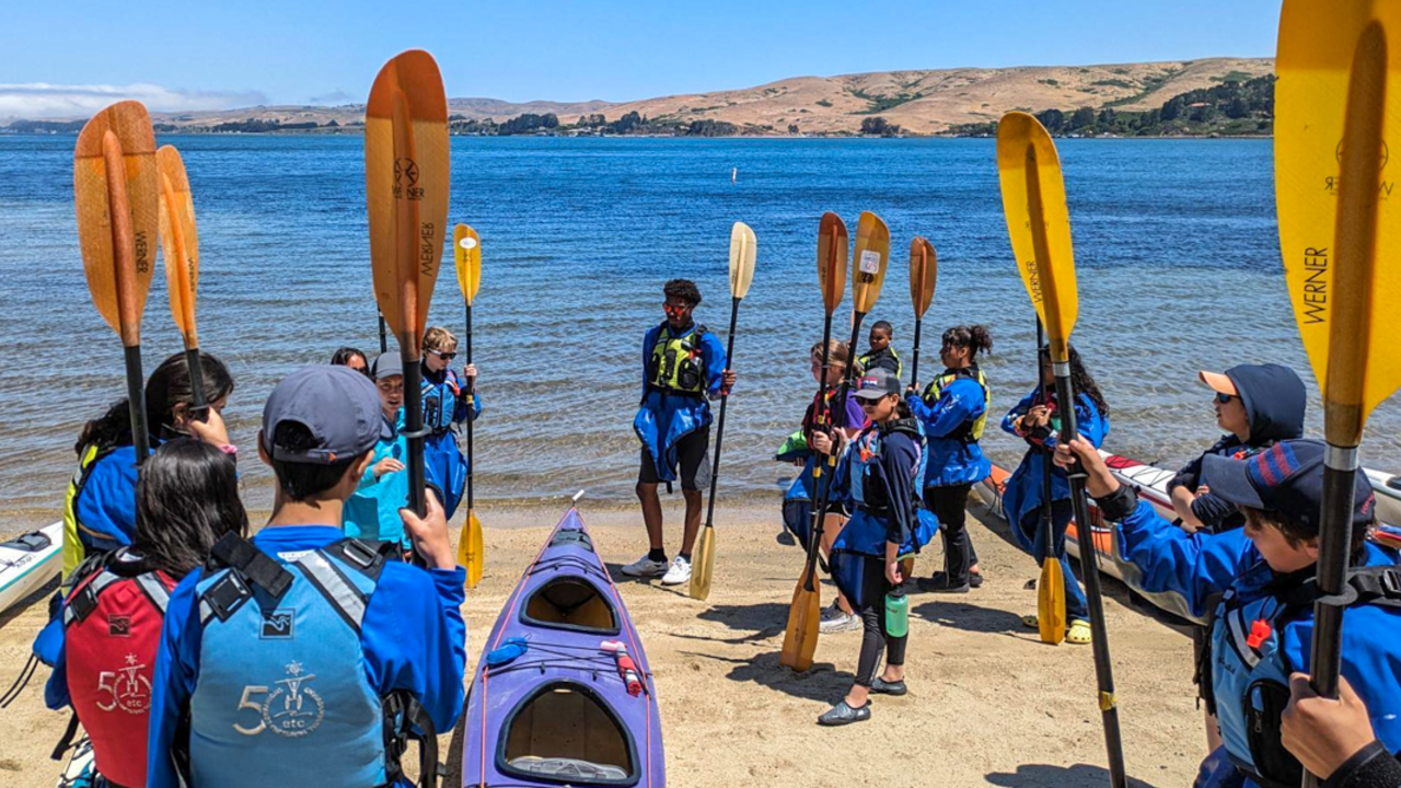 Crissy Field Center youth participants on the sunny shore of Hearts Desire Beach with kayaks and paddles