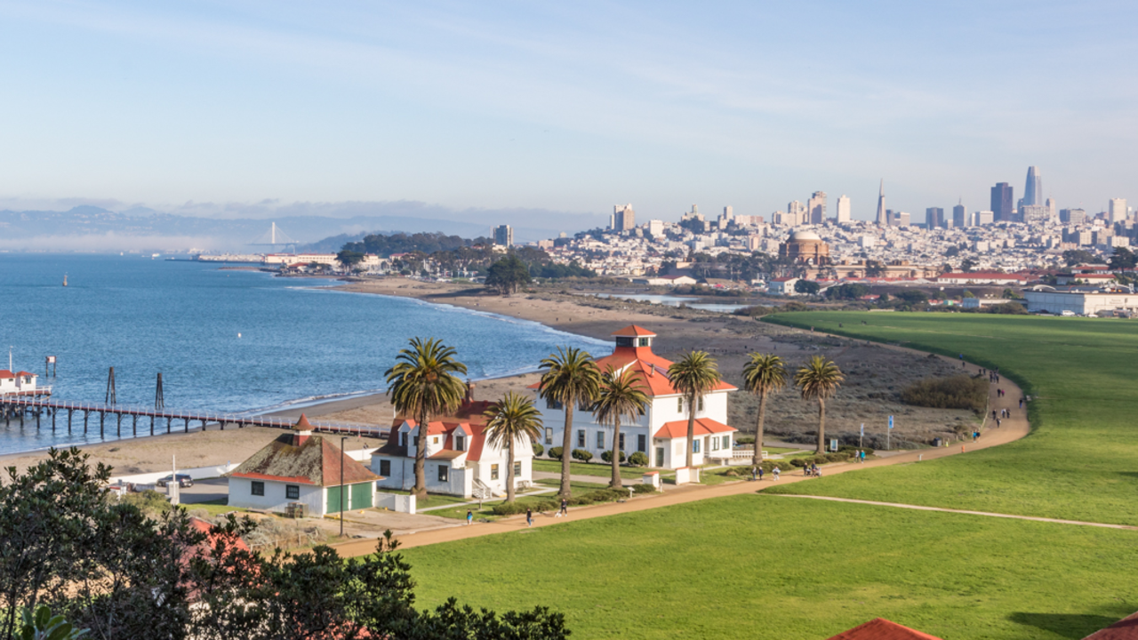 Aerial view of Crissy Field, buildings, the Warming Hut, palm trees, the golden gate, bay, and San Francisco city skyline.