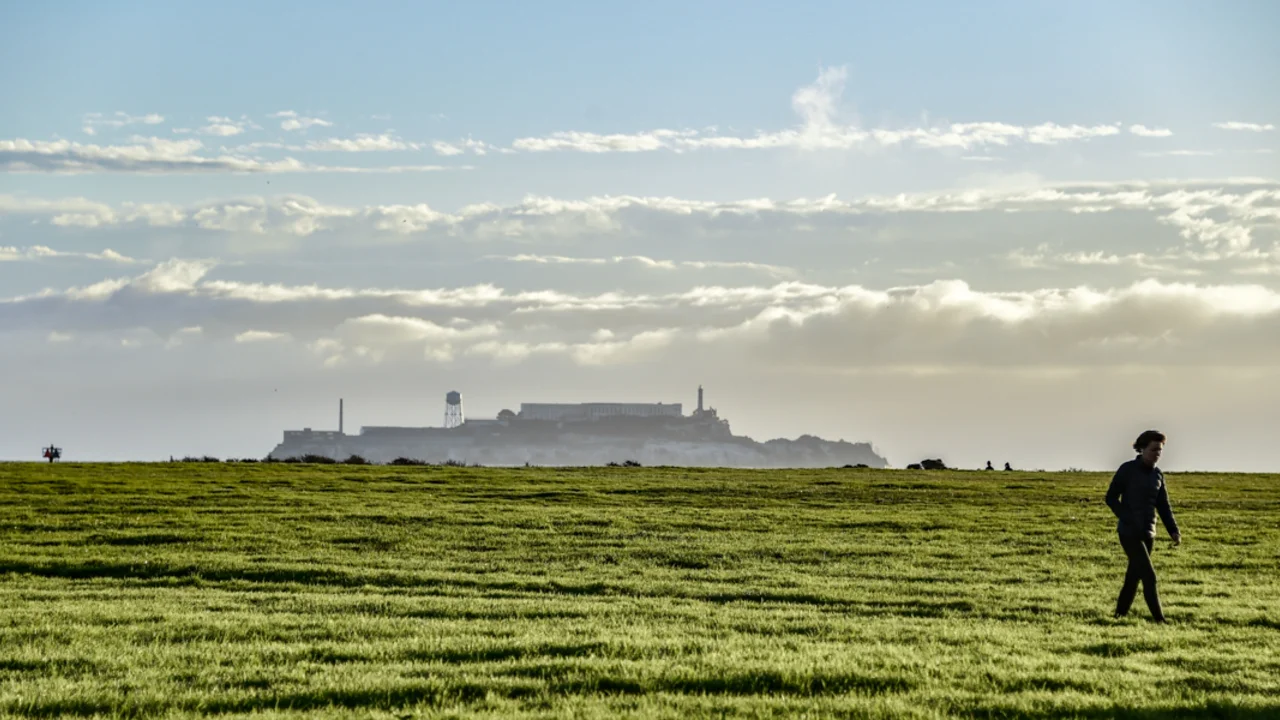 A view of Alcatraz from Crissy Field. A park visitor walks across the green grass under the blue sky.