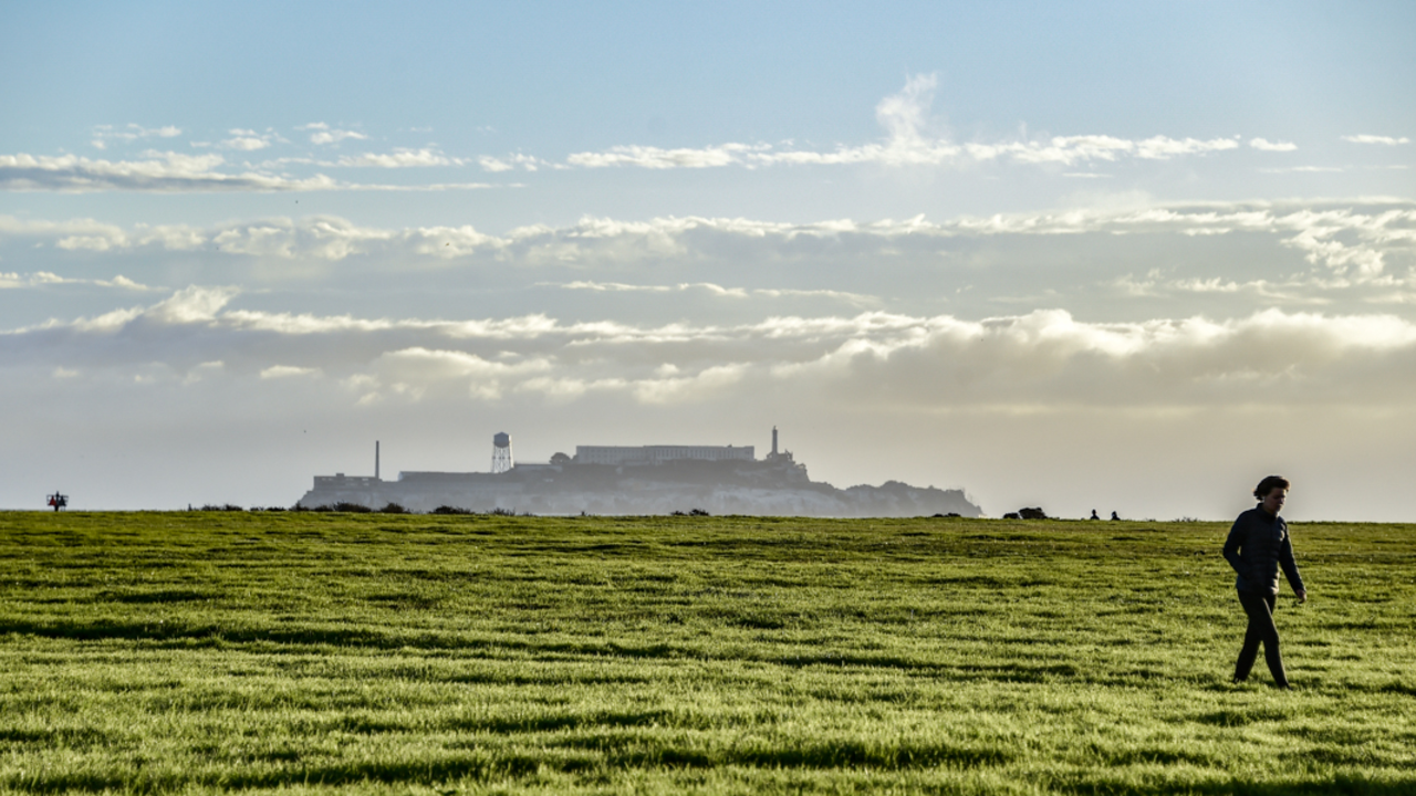 A View of Alcatraz from Crissy Field A view of Alcatraz from Crissy Field. A park visitor walks across the green grass under the blue sky.
