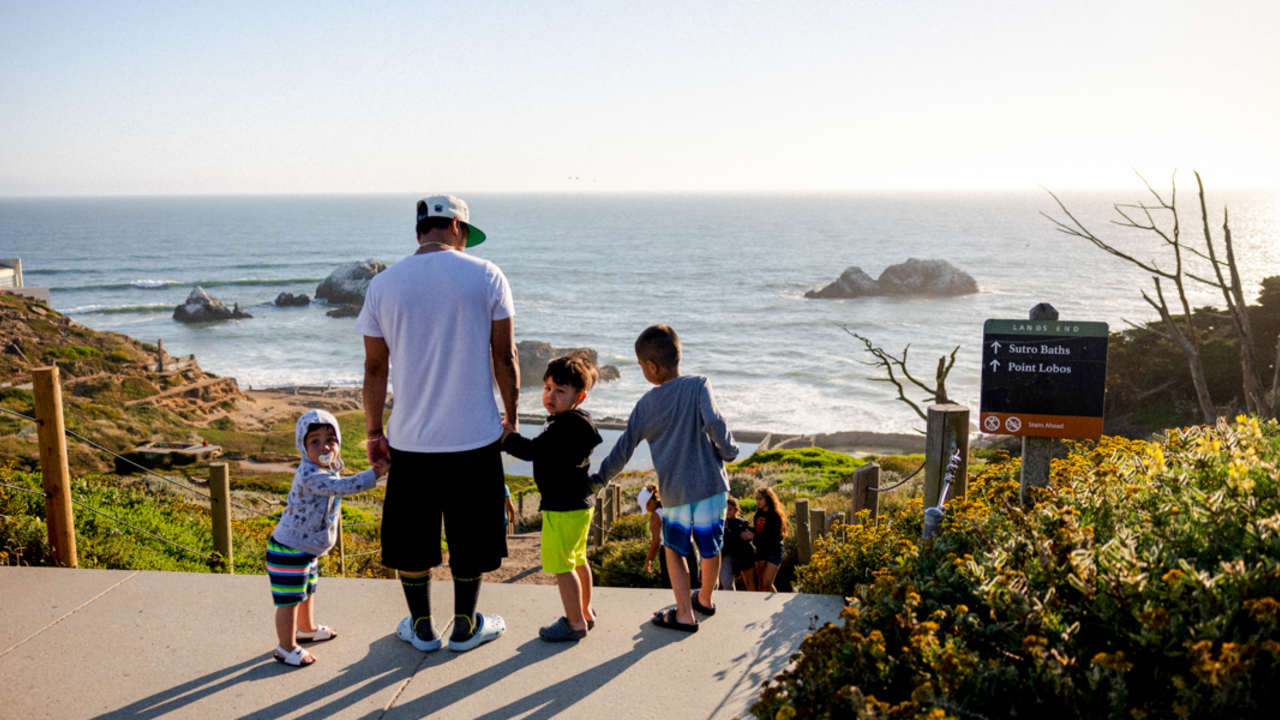 Lands End Visitors A family looks out over Sutro Baths at Lands End, with the sun setting over the pacific ocean.