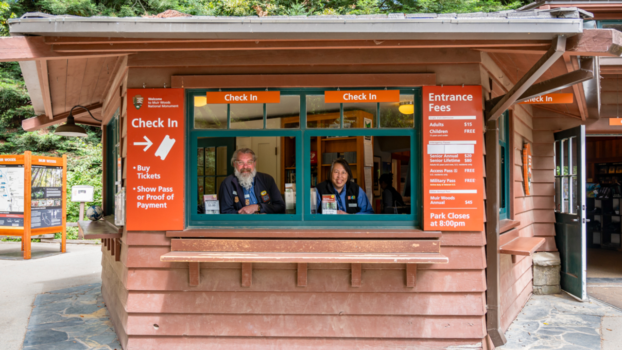 Parks Conservancy employees working at Muir Woods. Two smiling Parks Conservancy staff members stationed in the Muir Woods check in kiosk windows.
