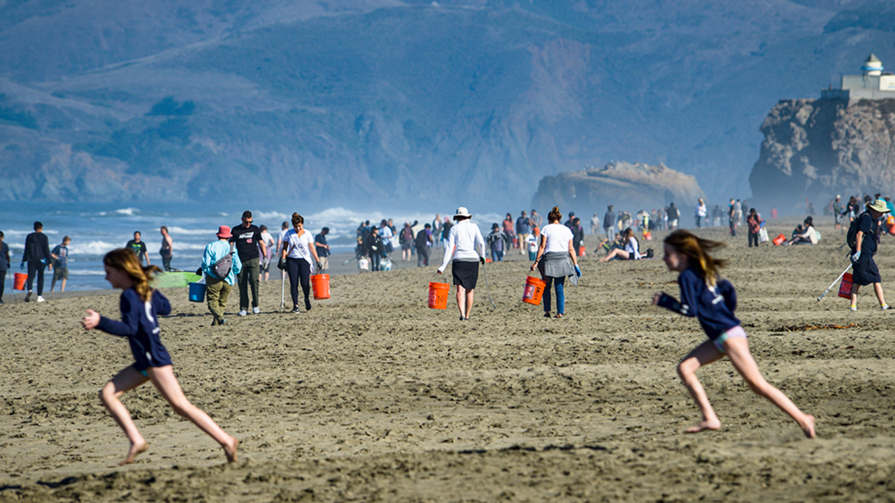 Two girls run in foreground of a wide shot of a crowded Ocean Beach.