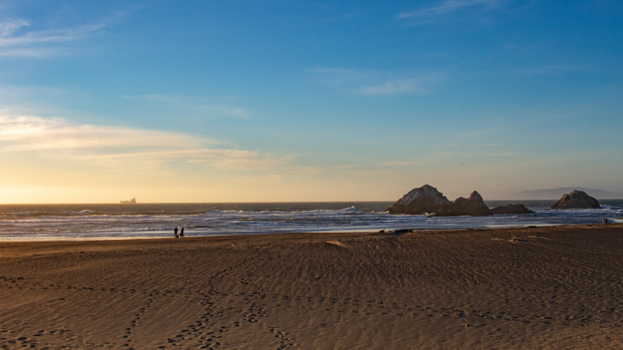 An evening view at Ocean Beach The open expanse of Ocean Beach shows an evening view of sandy shore, park visitors, ocean waves, and a commercial ship upon the horizon.