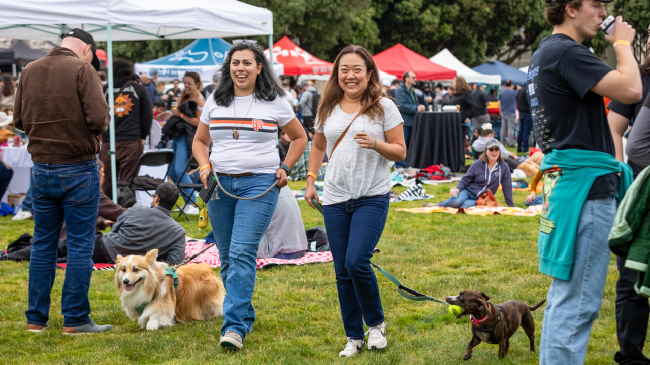 Two women walk their dogs amongst the lawn at Brewfest 2024 in the Presidio