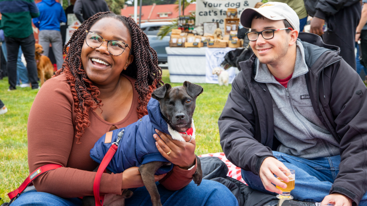 A smiling couple sits with their dog at the Parks4All: Brewfest in the Presidio
