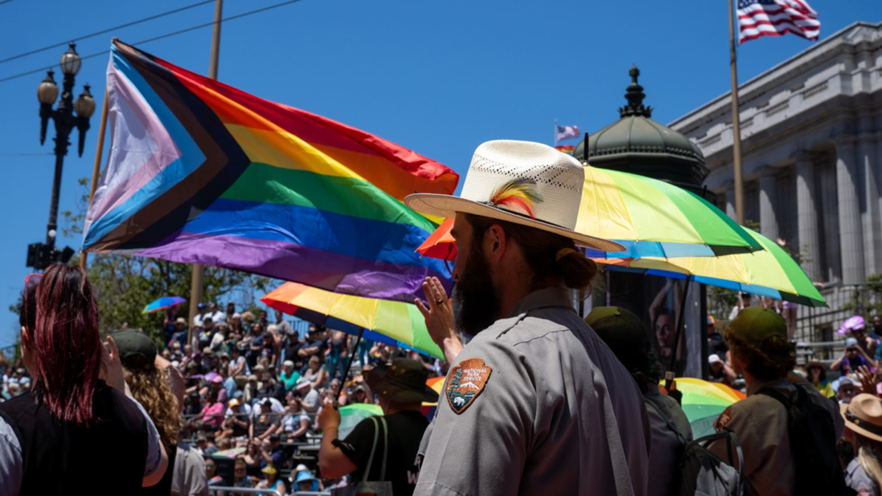 The side of a ranger's hat with a rainbow feather as he marches in the Pride Parade. Rainbow flags and umbrellas billow in the background.