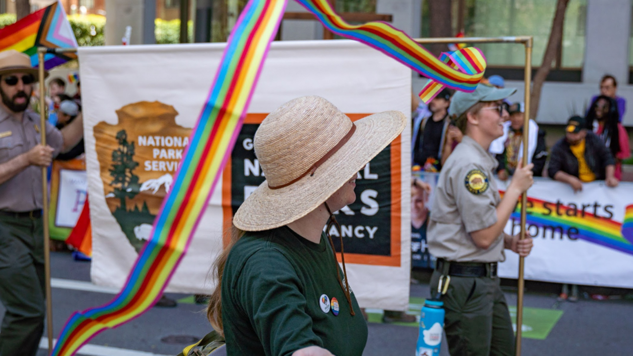 A ranger in a straw hat swirls a rainbow ribbin, while two other rangers hold a Golden Gate National Parks banner in the background.