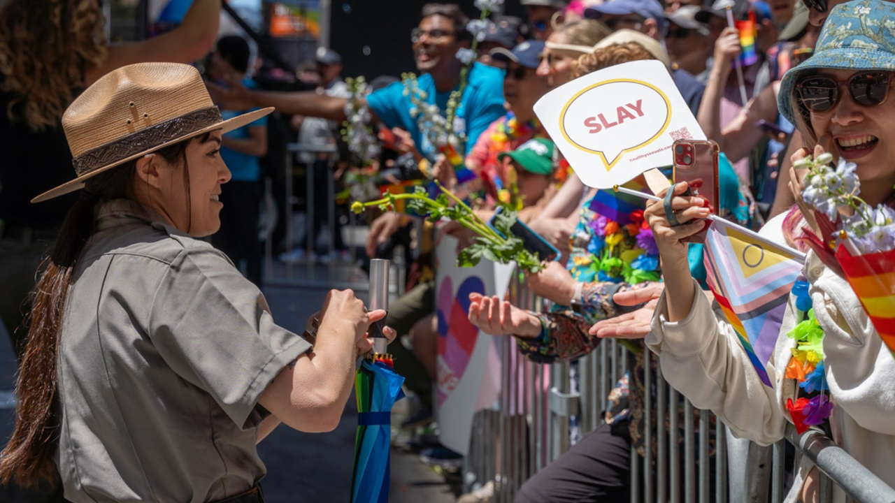 A ranger interacts with an excited crowd that's holding rainbow flags, cellphones, and a sign that says "SLAY".