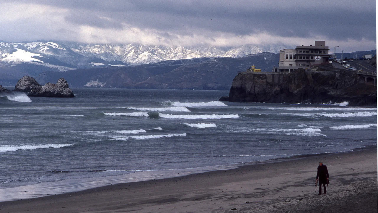 Person walking along Ocean Beach, in front of Cliff House, with snowy Marin Headlands in Background
