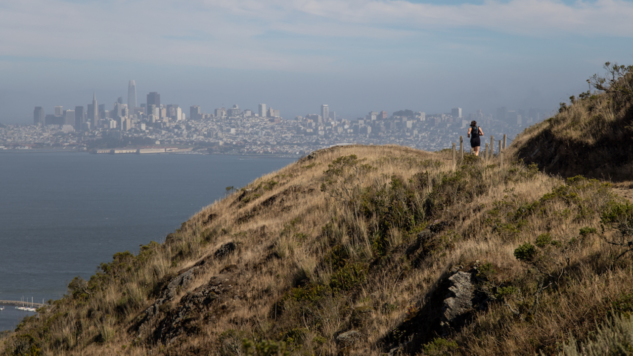 A park visitor runs on the SCA Trail in the Marin Headlands with the San Francisco skyline in background.
