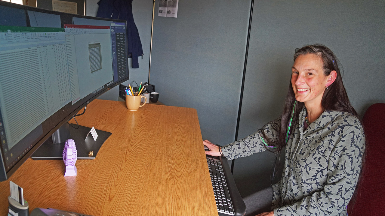 Lizzy at her office in the Marin Headlands Lizzy Edson smiles behind a large curved computer monitor.