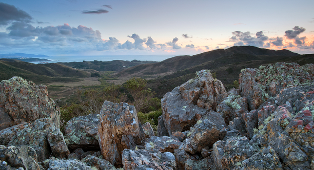 Scenic landscape view of Gerbode Valley in the Marin Headlands. Rocky boulders in the foreground frame the green expanse of the valley and mountains, and a pleasant sky with light clouds.