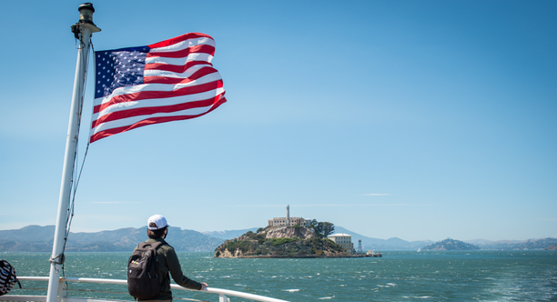 A park visitor looks toward Alcatraz Island from the ferry. An American flag waves in the breeze as they cruise over bay waters.