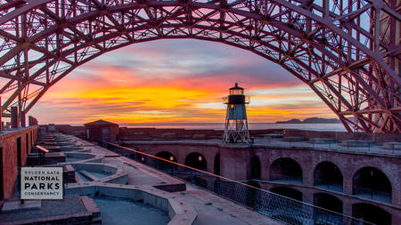 Lighthouse at Fort Point during sunset with Golden Gate Bridge arching above