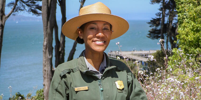 National Park Ranger Alanna Smith, a young woman, stands in the Presidio with the Pacific Ocean visible through the trees behind her.