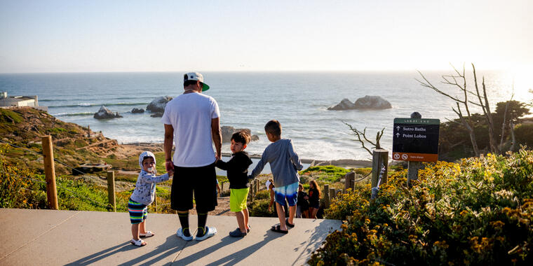 A family looks out over Sutro Baths at Lands End, with the sun setting over the pacific ocean.