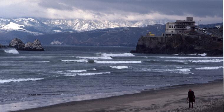 Person walking along Ocean Beach, in front of Cliff House, with snowy Marin Headlands in Background