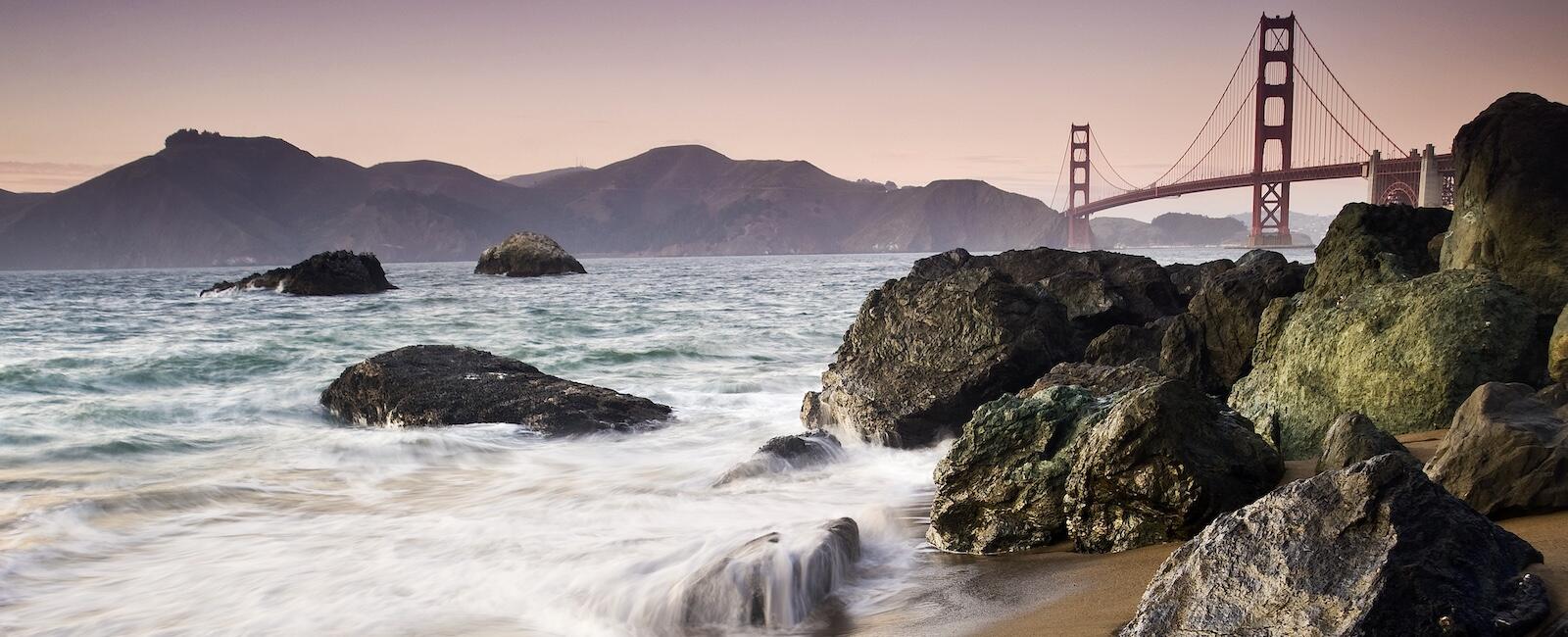 Waves crash over rocks at Marshall's Beach with Golden Gate Bridge in background.